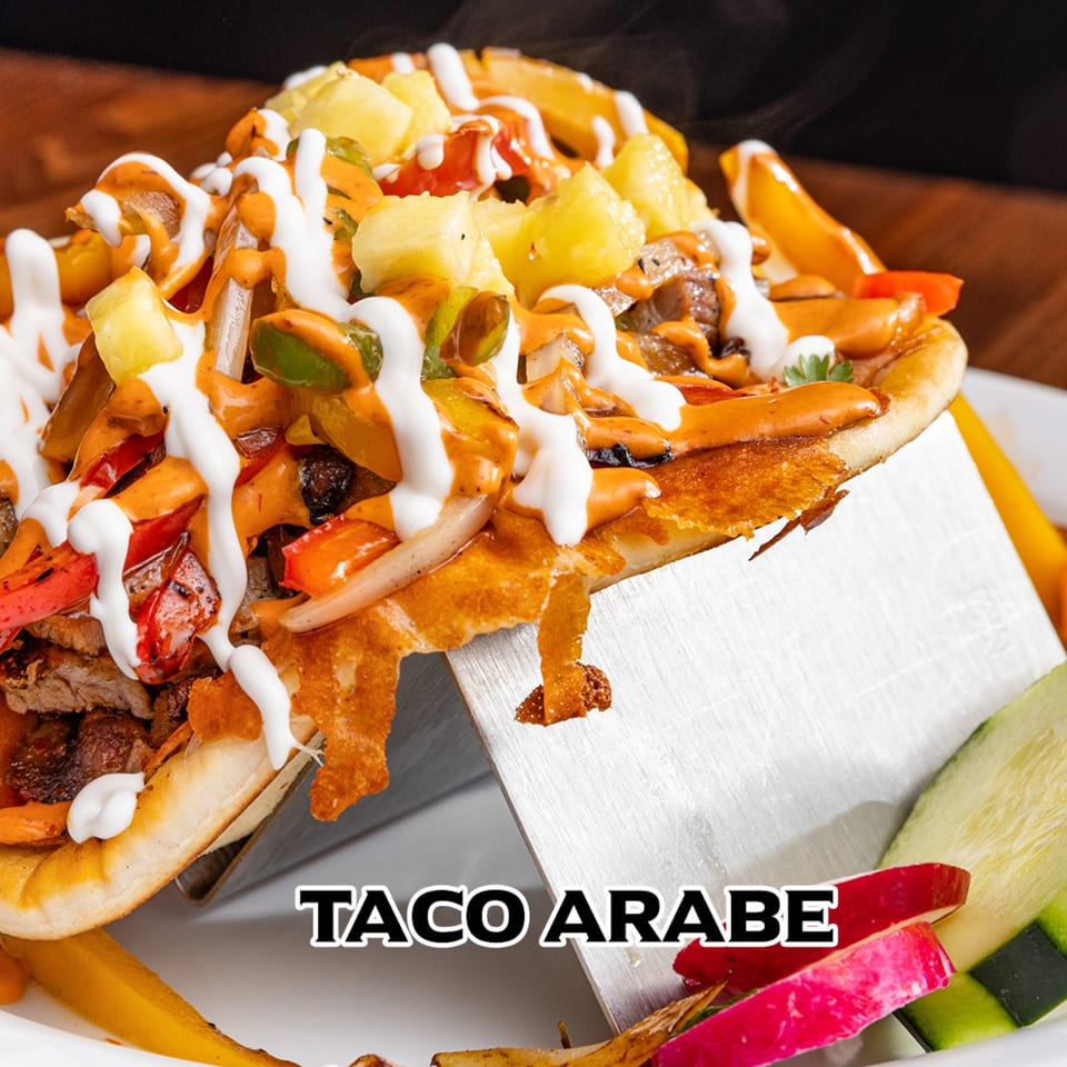 A House of Tacos Grill Taco Arabe, filled with meat, vegetables, pineapple, and sauces, on a metal stand.