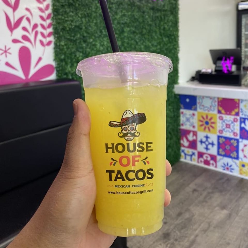 Hand holding a House of Tacos Grill drink with a colorful Mexican-themed background.