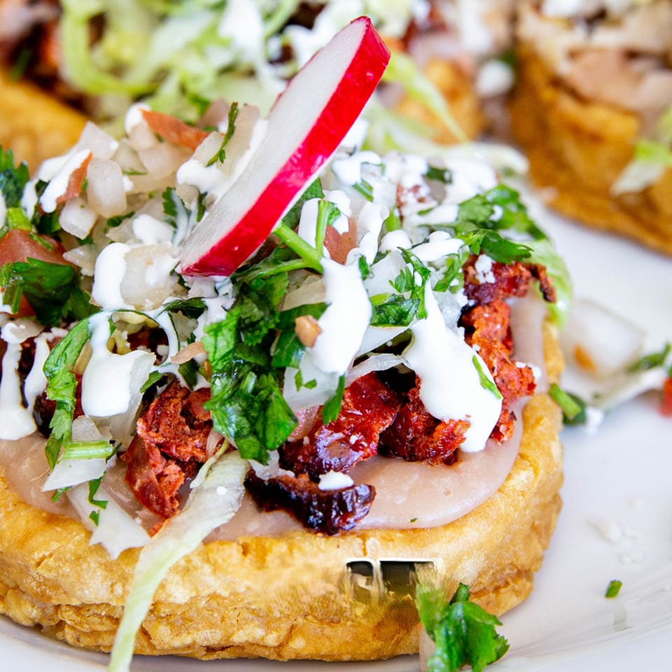 Close-up of a House of Tacos Grill sope, topped with meat, beans, lettuce, salsa, and crema.