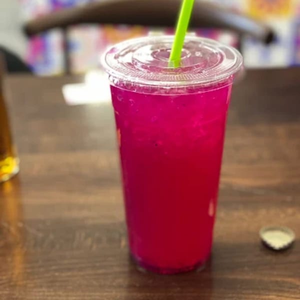 Bright pink drink with a green straw in a clear plastic cup on a wooden table.