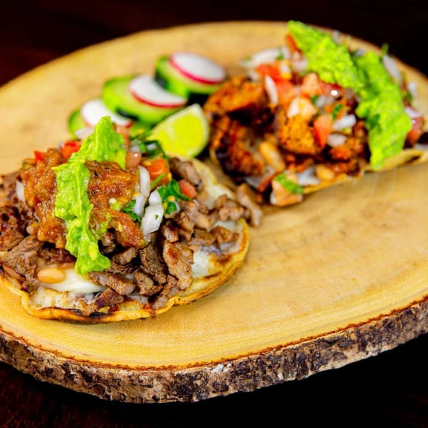 Two Mexican sopes with meat, guacamole, and pico de gallo, served on a wooden board.