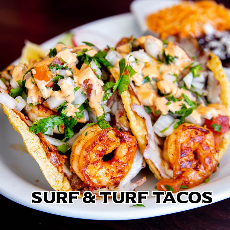 Close-up of two surf and turf tacos with shrimp, steak, salsa, and sauce on a white plate.