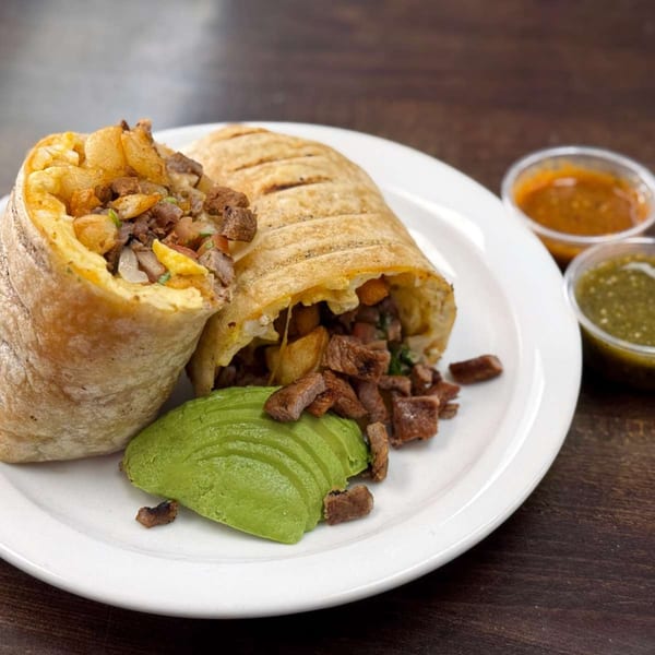 A grilled burrito, cut in half, revealing steak and potatoes, served with avocado and two sauces.